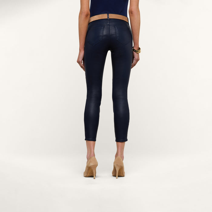 Wearing the Margot Coated Cropped Skinny Jean, which features a contoured waistband and a tapered leg in navy blue, along with a dark top and beige high-heeled shoes, a person stands facing away against a plain white background.