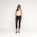 A woman wearing sunglasses, a white sleeveless top, the Margot Cropped Skinny Jean—black high-rise skinny jeans made from stretch denim with a contoured waistband for a flattering fit—a black belt with a gold buckle, and black heels stands against a plain white background.
