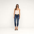 A woman wearing sunglasses, a white sleeveless top, Margot Cropped Skinny Jeans in dark blue premium stretch denim, a black belt, and black heels stands against a plain white background.