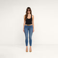 A woman wearing sunglasses, a black tank top, the Margot Cropped Skinny Jean in a vintage blue wash, a tan belt, and nude heels stands against a plain white background.