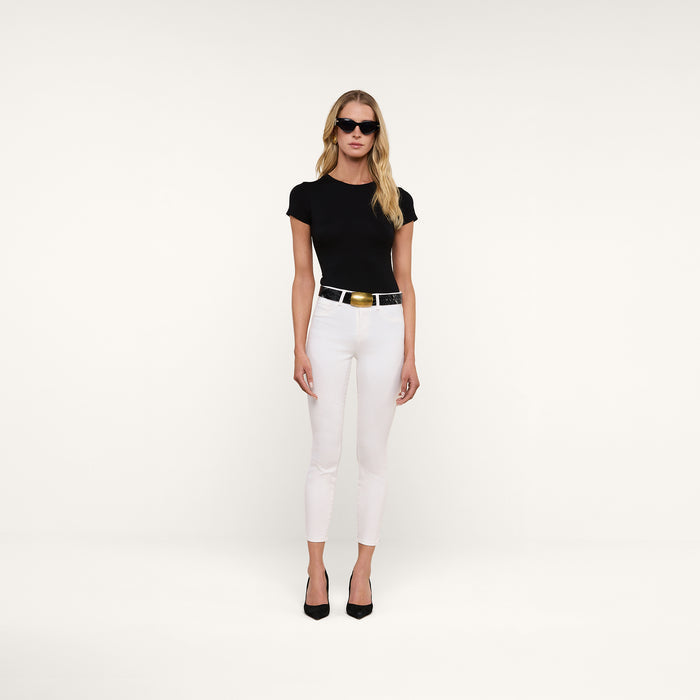 A woman wearing black sunglasses, a black t-shirt, a black belt with a gold buckle, the Margot Cropped Skinny Jean in white, and black heels stands against a plain white background.