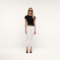 A woman wearing black sunglasses, a black t-shirt, a black belt with a gold buckle, the Margot Cropped Skinny Jean in white, and black heels stands against a plain white background.