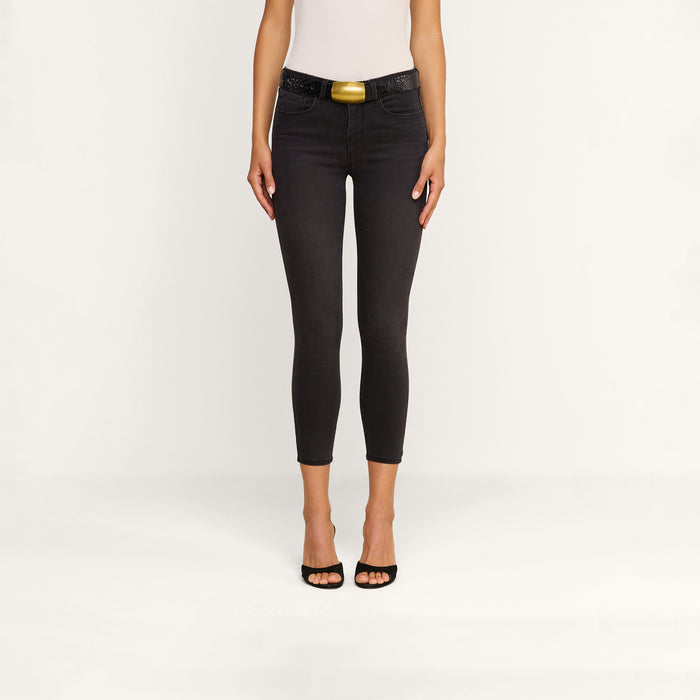 A woman stands against a plain background wearing the Margot Cropped Skinny Jean in black, featuring a contoured high-rise waistband and styled with a belt. She pairs the jeans with a white sleeveless top and black open-toe heels. Her arms are by her sides, and her head is not visible in the image.
