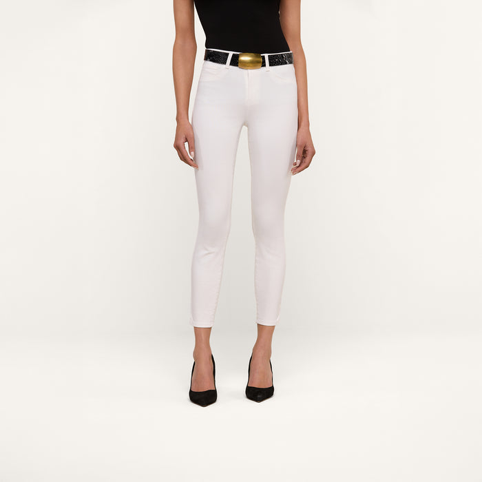 A person wearing a black sleeveless top, Margot Cropped Skinny Jeans in white with a shiny black belt and gold buckle, and black high heels stands against a plain white background.