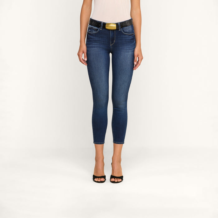A woman stands against a plain background, wearing the Margot Cropped Skinny Jean—crafted from premium stretch denim with sustainable recycled fiber—paired with a black belt featuring a gold buckle and black open-toe heels. Only her body from the shoulders down is visible.