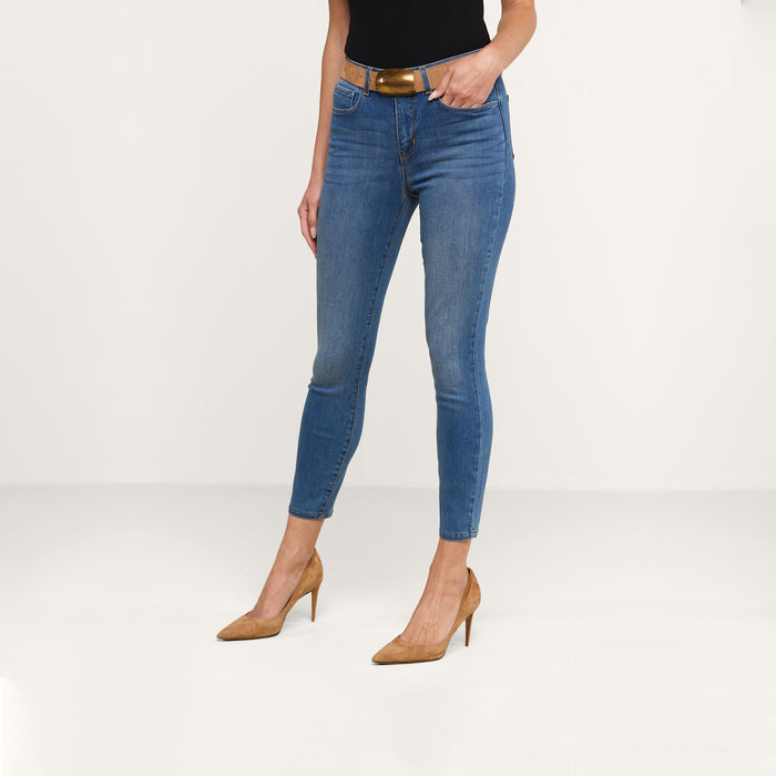 A person wearing the Margot Cropped Skinny Jean in blue stretch denim, paired with a black top, brown belt, and tan high-heeled shoes, stands against a plain white background.