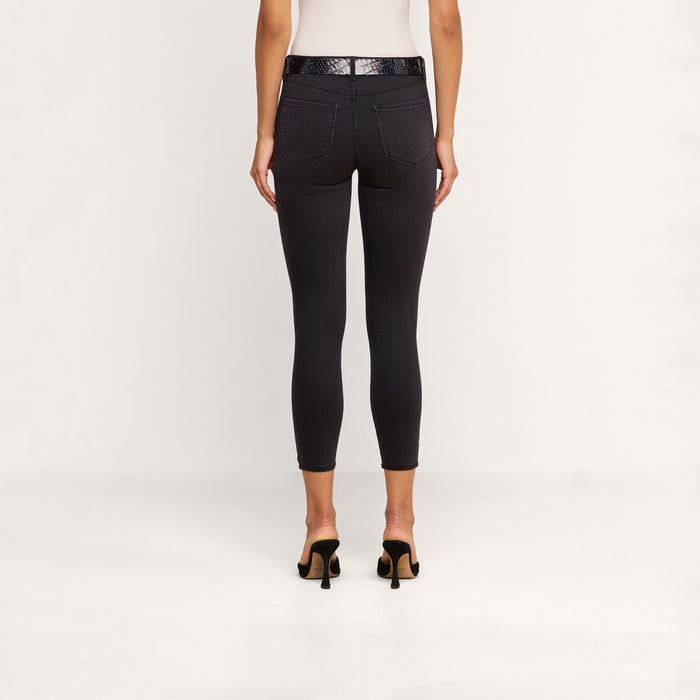 A woman stands facing away, wearing a white top and the Margot Cropped Skinny Jean—black high-rise skinny jeans with a contoured waistband—styled with a black belt and black heeled sandals against a plain light background.