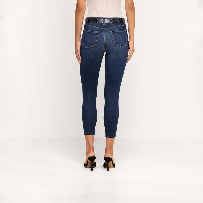 A person stands facing away against a plain white background, wearing a white sleeveless top, a black belt, black heeled sandals, and dark blue Margot Cropped Skinny Jeans crafted from premium stretch denim with back pockets.