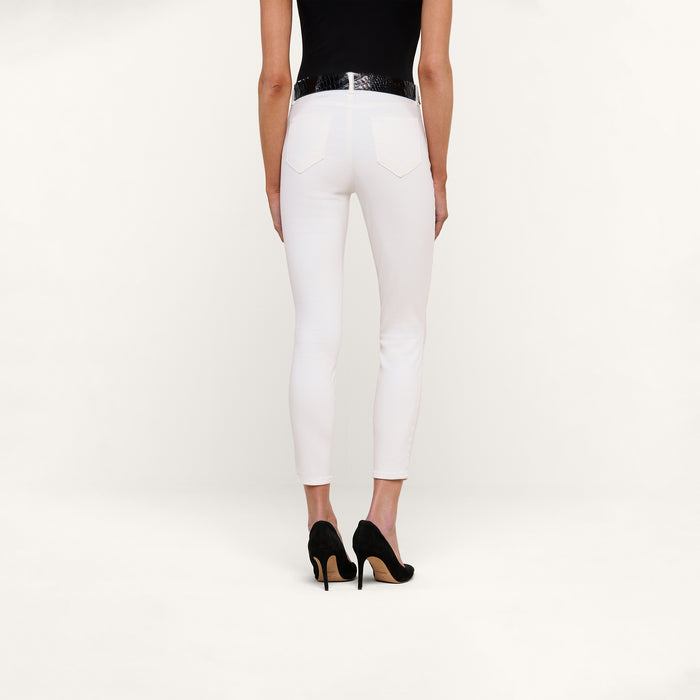 A person stands against a plain white background, shown from the back, wearing a black top and the Margot Cropped Skinny Jean in high-rise skinny white stretch denim with back pockets, paired with a black belt and black high-heeled shoes.