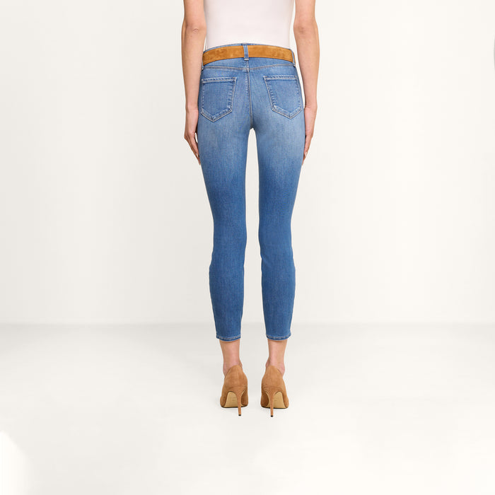 Person standing facing away, wearing the Margot Cropped Skinny Jean in blue, crafted from premium stretch denim, paired with a white top, a brown belt, and tan high-heeled shoes against a plain white background.