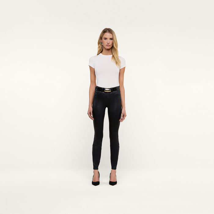 A woman stands facing forward against a plain white background, arms relaxed at her sides, wearing a fitted white short-sleeve top, black heels, a belt, and the Marguerite Coated Skinny Jean in high-waisted black coated denim.