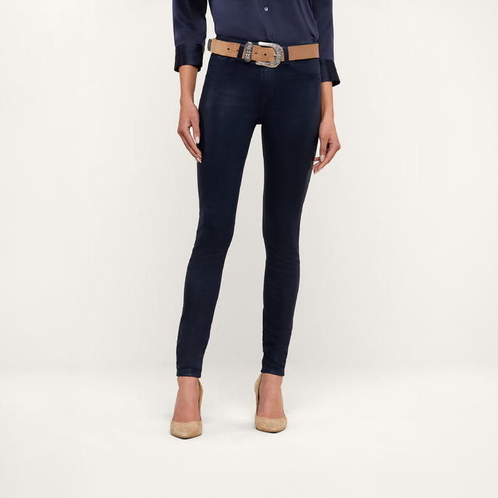 A person stands against a plain background, wearing a navy blouse, the Marguerite Coated Skinny Jean in navy with a tan belt, and tan high-heeled shoes. The photo is cropped at the shoulders.
