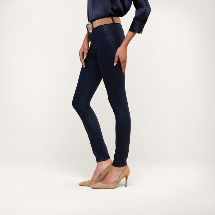 A person wearing the Marguerite Coated Skinny Jean in navy with a tapered leg, paired with a matching navy blouse, a tan belt, and tan high heels stands against a plain white background.
