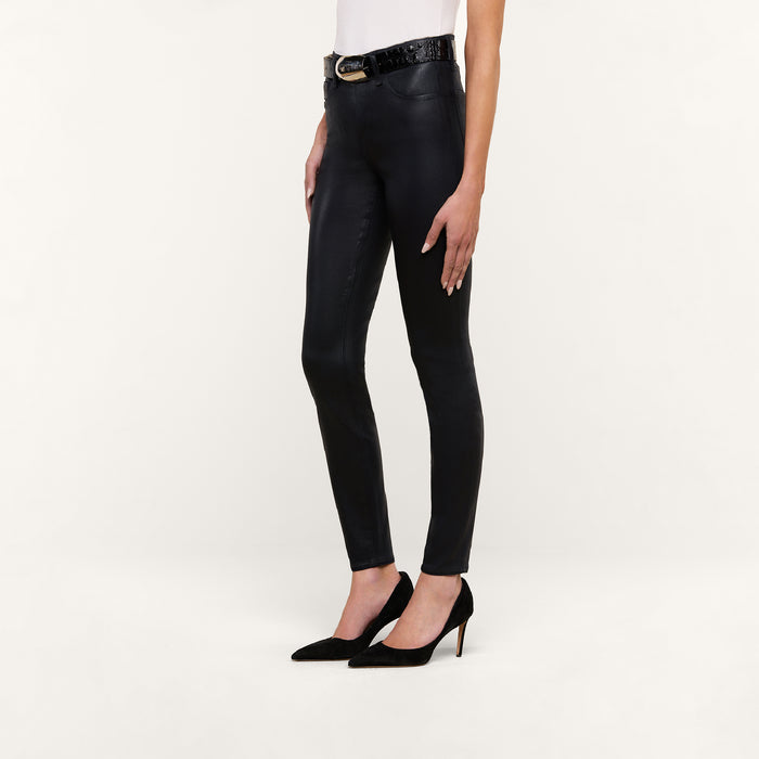 Wearing the Marguerite Coated Skinny Jean in black, a person styled a white top, a black belt, and black high-heeled shoes while standing against a plain white background.