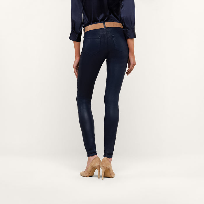 A person stands facing away, wearing a navy blouse and the Marguerite Coated Skinny Jean in navy with a tapered leg, paired with beige high-heeled shoes against a plain white background.