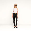A woman with long blonde hair wears sunglasses, a white t-shirt, the Marguerite Skinny Jean in black crafted from high-recovery fabric, a black belt with a gold buckle, and black heels, standing against a plain white background.