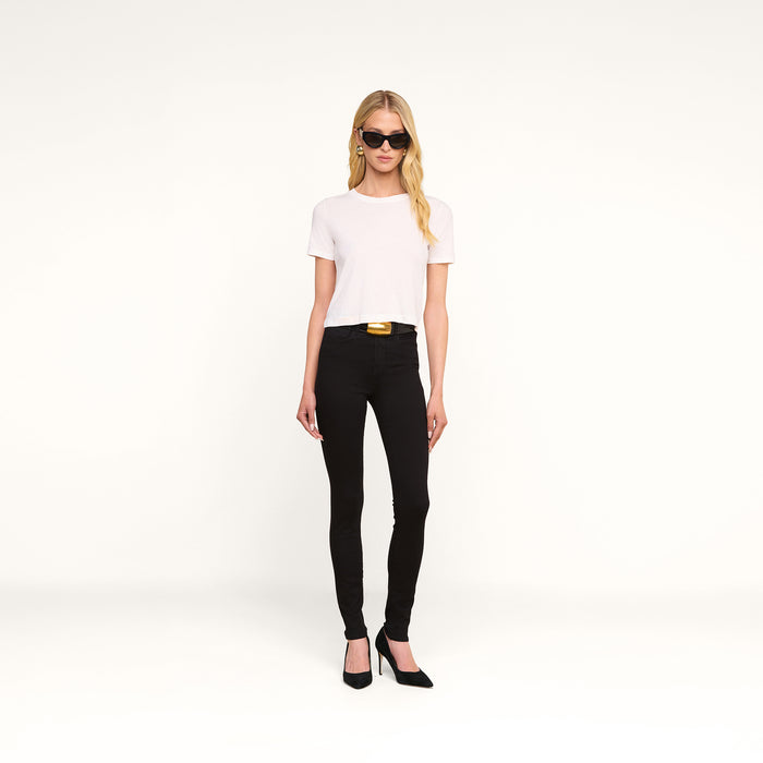 A woman with long blonde hair wears sunglasses, a white t-shirt, the Marguerite Skinny Jean in black crafted from high-recovery fabric, a black belt with a gold buckle, and black heels, standing against a plain white background.