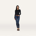 A woman stands against a plain white background wearing a black long-sleeve top, the Marguerite Skinny Jean in blue crafted from premium stretch denim, a brown belt, and black heeled sandals.