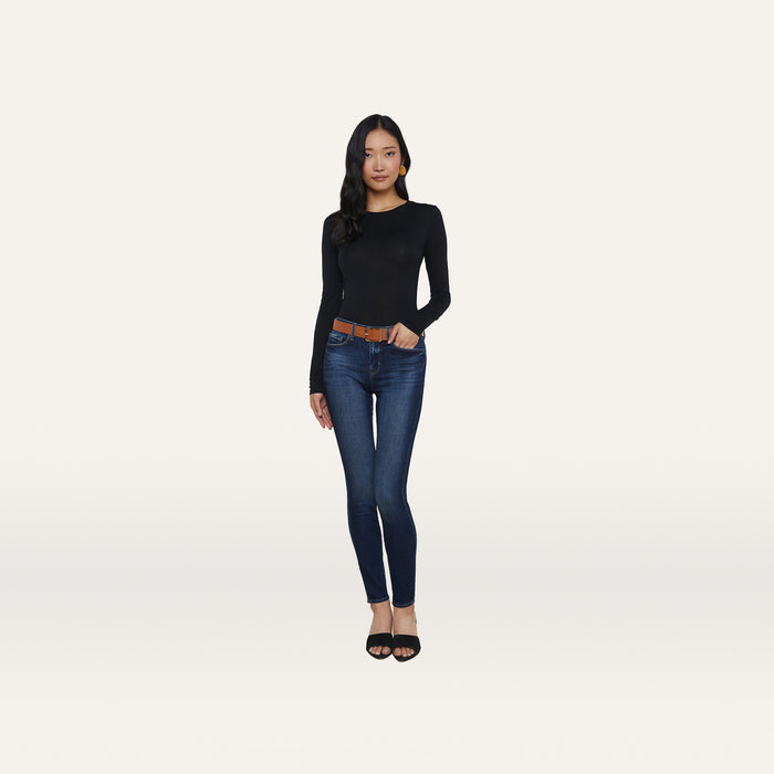 A woman stands against a plain white background wearing a black long-sleeve top, the Marguerite Skinny Jean in blue crafted from premium stretch denim, a brown belt, and black heeled sandals.