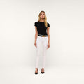 A woman stands against a plain white background wearing a black short-sleeve top, the Marguerite Skinny Jean crafted from high-recovery white denim with a shiny silver belt, and black heels.