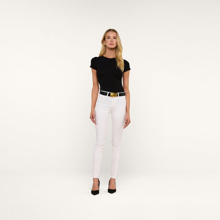 A woman stands against a plain white background wearing a black short-sleeve top, the Marguerite Skinny Jean crafted from high-recovery white denim with a shiny silver belt, and black heels.