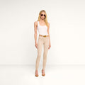 A woman wearing sunglasses, a white sleeveless top, the Marguerite Skinny Jean in beige high-recovery fabric, a brown belt, and brown heels stands against a plain white background.