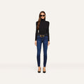 Wearing sunglasses, a black turtleneck sweater, black heels, and the Marguerite Skinny Jean with a belt, a woman stands against a plain, light background.