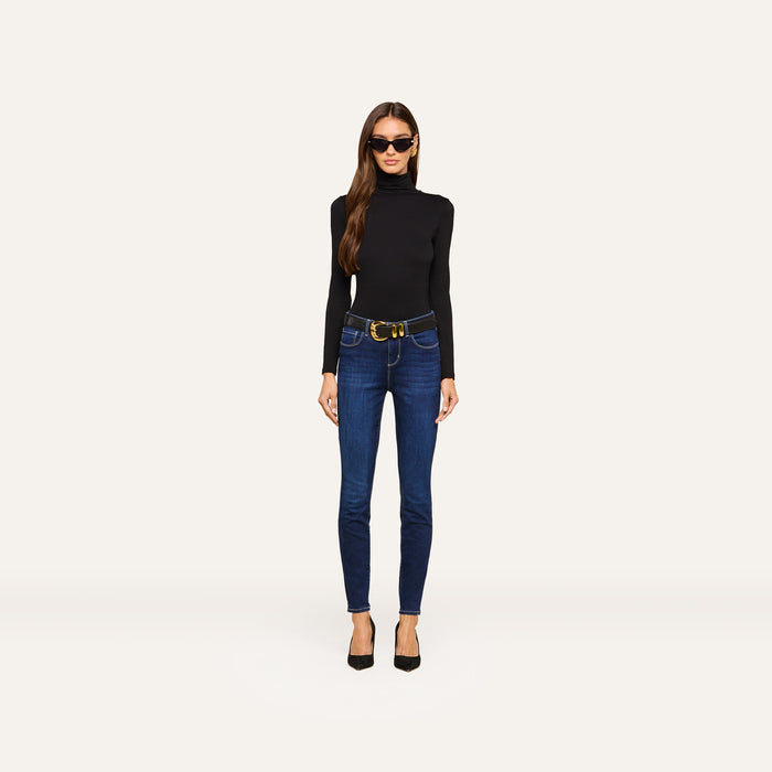 Wearing sunglasses, a black turtleneck sweater, black heels, and the Marguerite Skinny Jean with a belt, a woman stands against a plain, light background.