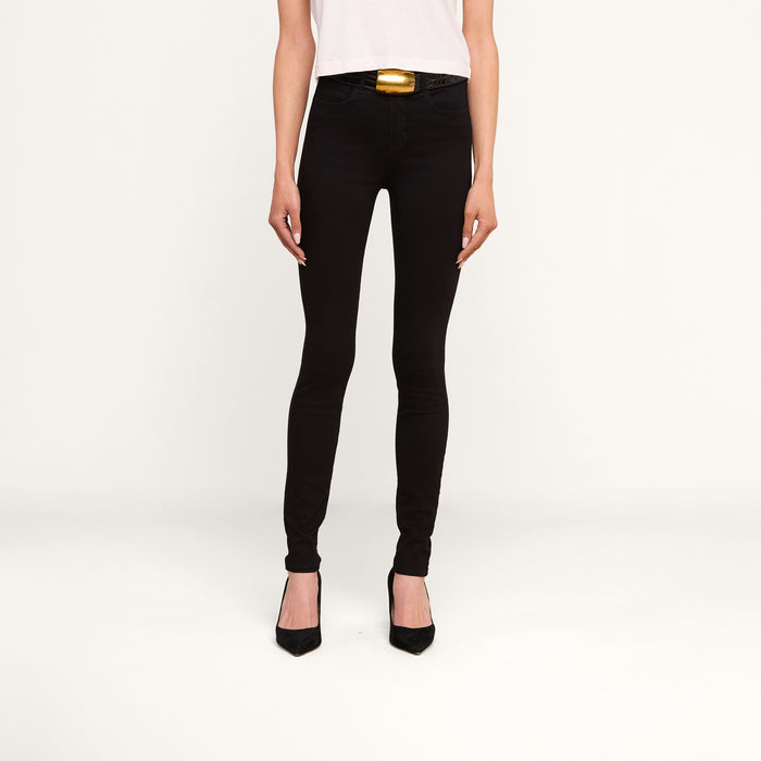 A person wearing the Marguerite Skinny Jean in black high-recovery fabric, paired with a white shirt and black pointed-toe heels, stands against a plain white background. Only the lower half of the body is visible.