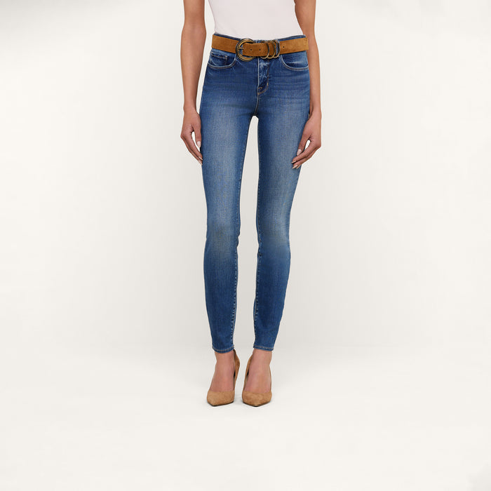 A woman is shown from shoulders to feet against a plain white background, wearing the Marguerite Skinny Jean in blue high rise stretch denim, paired with a brown belt, beige pointed heels, and a white top.