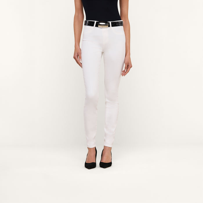 A woman stands against a plain background, wearing a black sleeveless top, the Marguerite Skinny Jean in white with a black belt, and black pointed-toe shoes. Only her body from the shoulders down is visible.