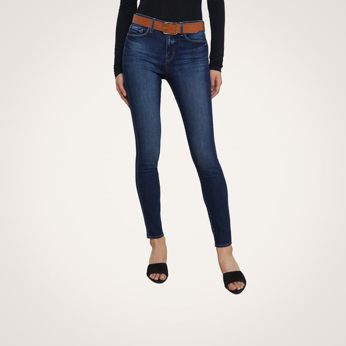 A person wearing the Marguerite Skinny Jean in dark blue—crafted from premium stretch denim—paired with a brown belt, a black long-sleeve top, and black open-toe shoes stands against a plain white background.