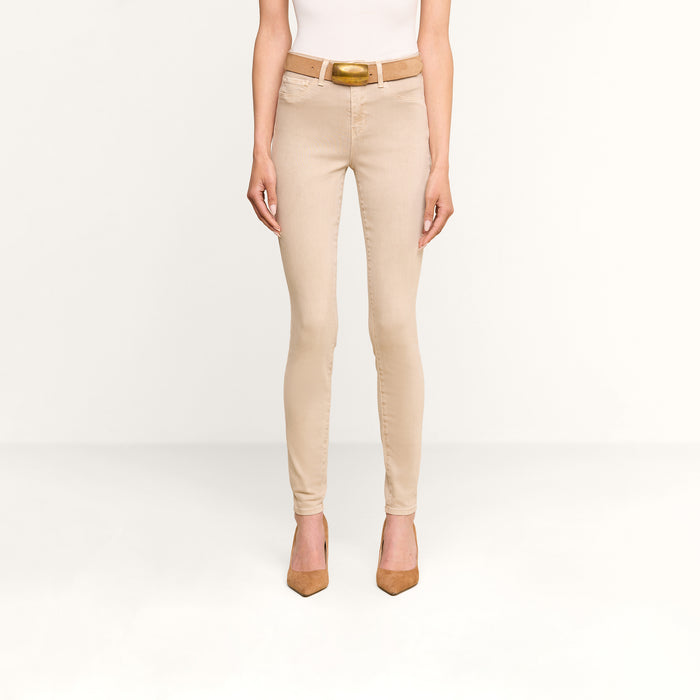 Wearing the Marguerite Skinny Jean in beige with a tapered leg, styled with a white fitted top, tan belt, and brown pointed high heels. The model is shown from the shoulders down against a plain white background.