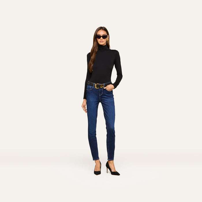 A woman wearing sunglasses, a black long-sleeve turtleneck, a belt, and black high heels stands against a plain light background. She completes her outfit with the Marguerite Skinny Jean—blue high rise skinny jeans crafted from stretch denim.