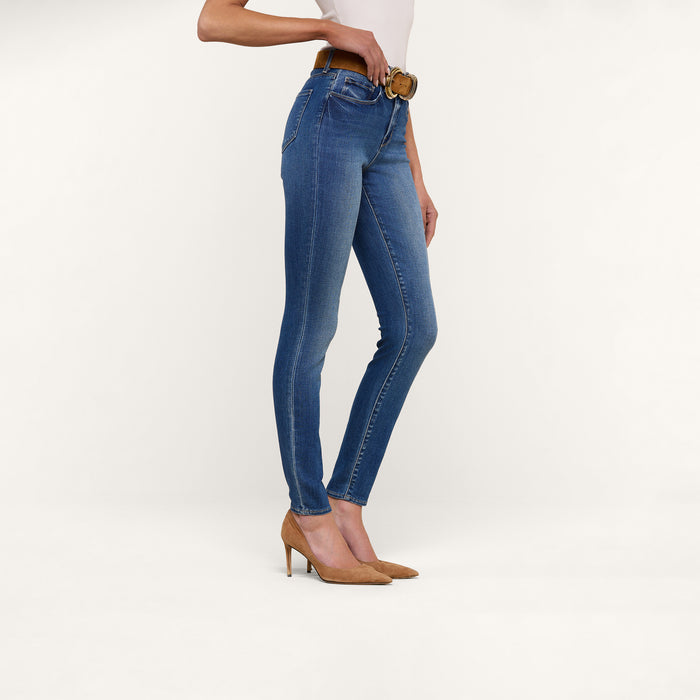 A person is shown from the shoulders down, standing against a plain white background, wearing the Marguerite Skinny Jean in blue stretch denim paired with a brown belt and tan high heels.