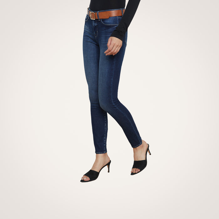 Wearing the Marguerite Skinny Jean crafted from premium stretch denim in dark blue, paired with a brown belt, a black long-sleeve top, and black heeled sandals, the person stands against a plain light background.