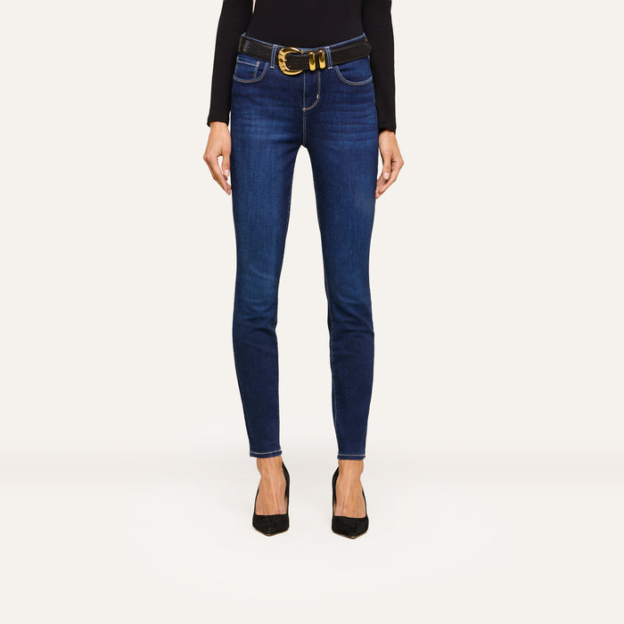 Person wearing dark blue skinny jeans, a black long-sleeve top, a wide black belt with a large gold buckle, and sleek black pointed-toe heels, standing against a plain white background.