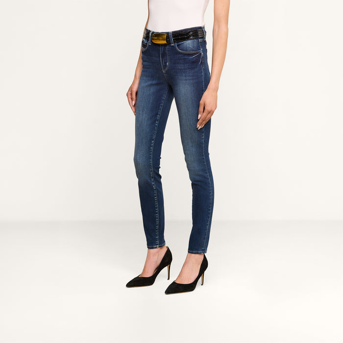 A person wearing the Marguerite Skinny Jean in premium stretch denim, paired with a white sleeveless top, a black belt, and black high-heeled shoes, stands against a plain light background.