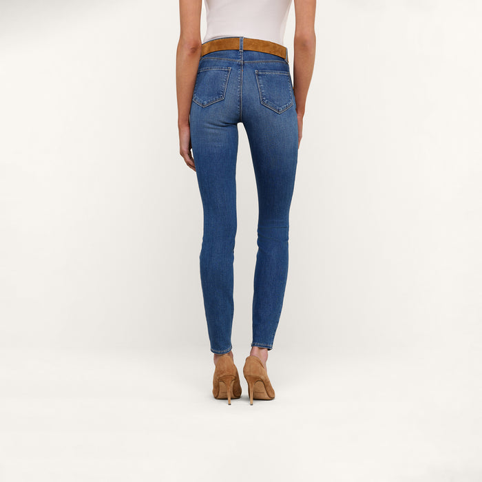 A person wearing the Marguerite Skinny Jean in blue stretch denim, paired with a brown belt and tan high heels, is standing against a plain white background, facing away from the camera.