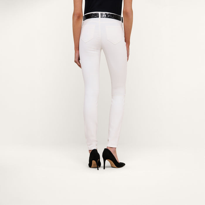Person standing against a plain background, shown from the waist down, wearing the Marguerite Skinny Jean in white high-recovery fabric, paired with a black top, a white belt, and black high-heeled shoes.