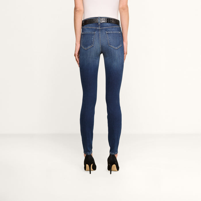 A person wearing a white top, the Marguerite Skinny Jean made from premium stretch denim, a black belt, and black high-heeled shoes is standing facing away from the camera against a plain white background.