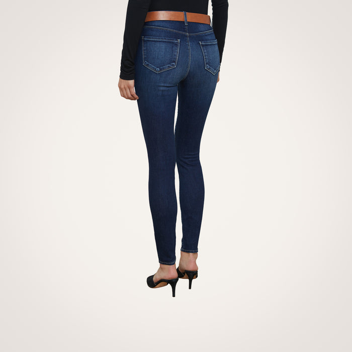 A person is shown from the back against a plain white background, wearing the Marguerite Skinny Jean in dark blue premium stretch denim, paired with a black long-sleeve top and black high heels.
