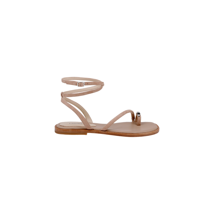 The Mariana Suede Sandal is a beige flat sandal featuring asymmetric straps, a slender ankle strap, a single toe strap with a metallic accent, and a minimal wooden sole, showcased in side profile against a white background.