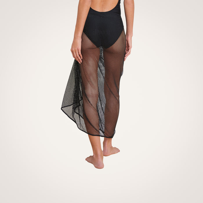 A woman stands barefoot against a plain white background, wearing a black swimsuit and the Maribel Mesh Pareo—a sheer rhinestone mesh skirt styled as a chic swimsuit cover-up.