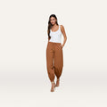 A woman wearing a white tank top, the Mariko Jogger in brown—crafted from water-repellent nylon—and high-heeled sandals stands against a plain white background, with one hand in her pocket.