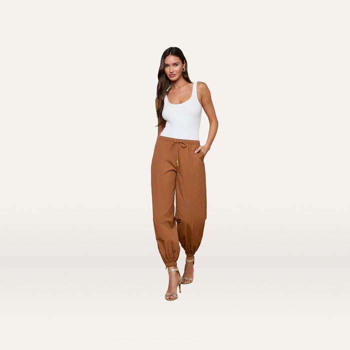 A woman wearing a white tank top, the Mariko Jogger in brown—crafted from water-repellent nylon—and high-heeled sandals stands against a plain white background, with one hand in her pocket.