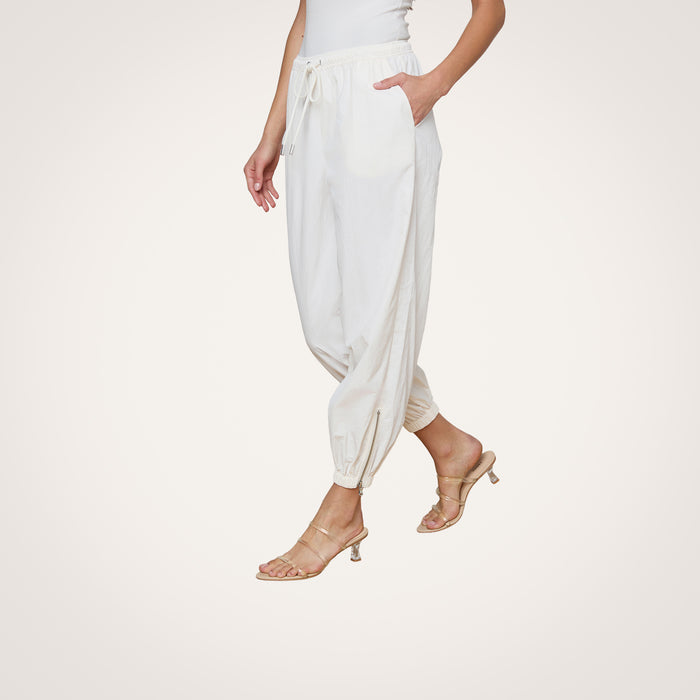 Wearing the Mariko Jogger in ivory—a modern silhouette crafted from water repellent nylon—paired with a white top and beige heeled sandals, a person stands against a plain, light background with one hand in their pocket and their face not visible.