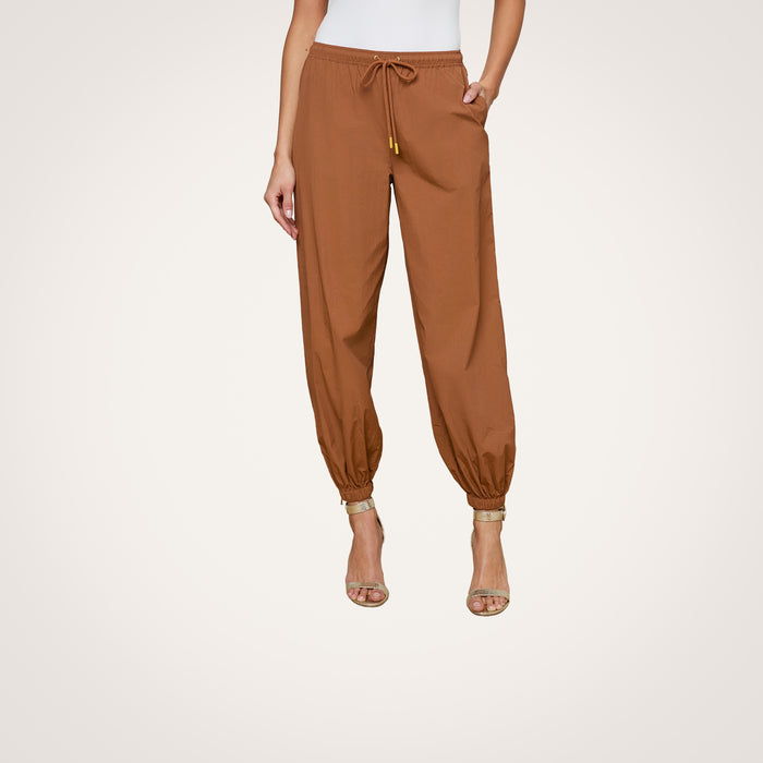 Wearing the Mariko Jogger—modern brown jogger pants crafted from water-repellant nylon with elastic cuffs—paired with beige heeled sandals, a person stands against a plain white background.