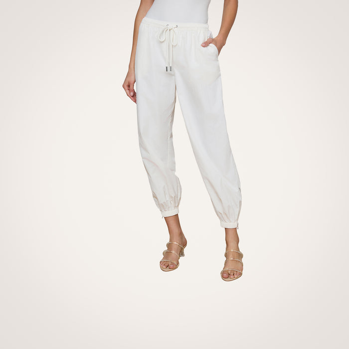 A woman stands against a light background with her hands in her pockets, wearing the Mariko Jogger in modern ivory, crafted from water-repellant nylon. Her beige heeled sandals are visible, while her upper body is mostly out of frame.
