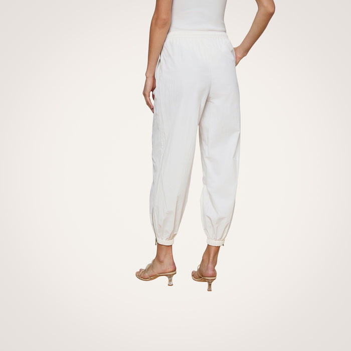 Someone is standing against a plain, light background, shown from the waist down and viewed from behind. They are wearing the Mariko Jogger—modern jogger-style pants in ivory crafted from water-repellent nylon—with heeled sandals.
